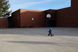 A toddler in a puffy blue jacket and a bright blue helmet rides a green balance bike in an empty basketball court dappled with long shadows; in the background is a two story brick building, the only visible windows are the glass in the entry way behind the only visible basketball hoop